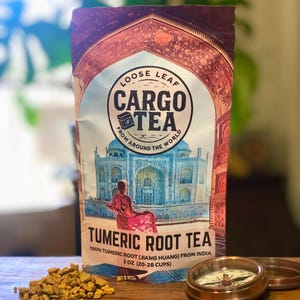 Puede incluir: Un paquete de té Cargo Tea Tumeric Root con una ilustración del Taj Mahal. El paquete incluye el texto "Loose Leaf Cargo Tea From Around The World" y "Tumeric Root Tea". También se ven una brújula y trozos de raíz de cúrcuma.