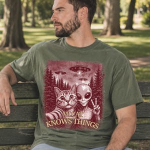 May include: Olive green t-shirt featuring a graphic of a cat and an alien posing together with UFOs in the background. The text "Mi Cat Knows Things" is printed below the image. The shirt is worn by a person.