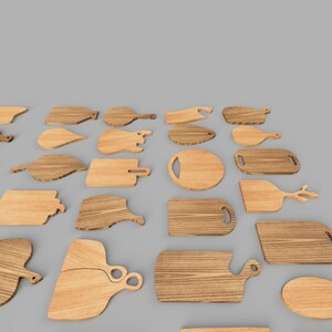 Op de afbeelding: Een verzameling verschillende houten snijplanken in verschillende vormen en maten. De planken zijn gemaakt van lichtbruin hout en hebben unieke handvatontwerpen, waaronder ronde, rechthoekige en peddelvormige opties. De planken zijn gerangschikt op een grijze ondergrond.