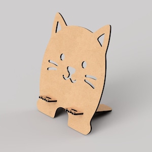 May include: A light brown cat-shaped phone stand made of wood. The stand features cut-out details for the eyes, nose, and whiskers. It has two small shelves at the bottom to hold a phone and a small support at the back to keep it upright.