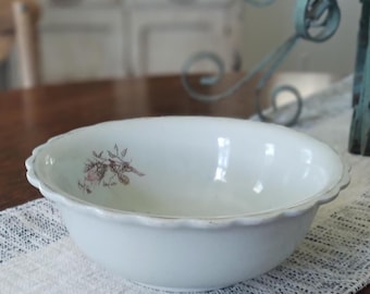 May include: A white ceramic bowl with a scalloped rim and a faded floral design. The bowl sits on a white and gray patterned table runner. The bowl is likely vintage or antique.