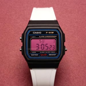 Puede incluir: Reloj digital Casio F-91W negro con correa blanca. La esfera del reloj muestra la hora, el día y la fecha en formato digital. El reloj tiene una esfera azul y negra con el logotipo de Casio.