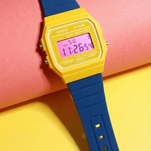 Puede incluir: Reloj digital Casio amarillo con correa azul. La esfera muestra la hora 11:26:54. El reloj tiene el texto "CASIO" y "WATER RESIST" impreso en la esfera.
