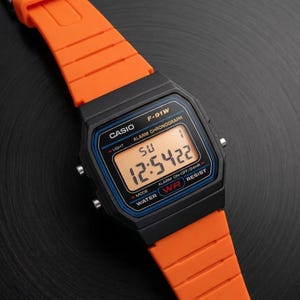 Op de afbeelding: Een zwarte Casio F-91W digitale horloge met een oranje band. De wijzerplaat toont de tijd, dag en datum. Het horloge heeft een zwarte kast met blauwe en witte accenten. De oranje band is gemaakt van flexibel materiaal.