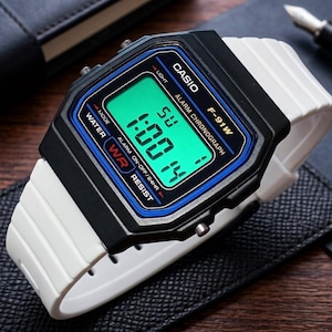 May include: A Casio F-91W digital watch featuring a black rectangular case with a blue border and a bright green digital display. The watch has a white resin strap and includes text on the face reading CASIO, F-91W, ALARM CHRONOGRAPH, and WATER RESIST.