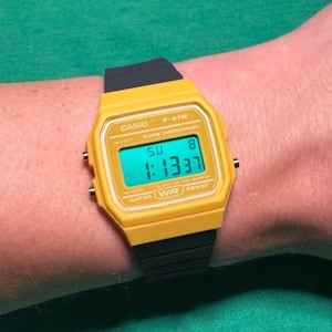 May include: A yellow Casio F-91W digital watch with a black strap is shown on a wrist. The watch face displays the time as 1:13:37 on a teal background. The watch also shows the day and date.