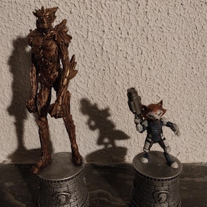 Peut inclure: Deux figurines détaillées de personnages de Gardiens de la Galaxie. L'une est une figurine marron en forme d'arbre, l'autre un personnage ressemblant à un raton laveur tenant une arme. Les deux sont sur des socles cylindriques avec un motif de briques.