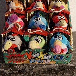 Puede incluir: Una caja expositora llena de juguetes de pájaros de peluche. Los pájaros están en compartimentos individuales, cada uno con un techo rojo. Los pájaros son de varios colores, incluyendo rojo, azul, verde y amarillo, con rasgos y expresiones de dibujos animados. La caja tiene el texto "Koo Koo Zoo Birds".