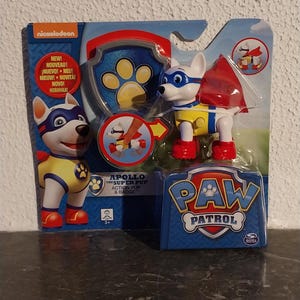 Op de afbeelding: Een Paw Patrol Apollo de Super Pup actiefiguur in een blauw en rood pakket. De figuur is wit met een blauw masker, geel vest en rode laarzen. De verpakking bevat het Paw Patrol logo en het Nickelodeon logo.