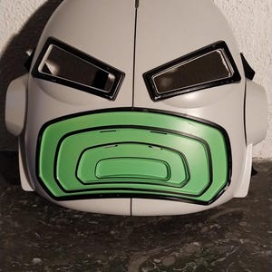 Könnte beinhalten: Eine grau-graue, futuristisch aussehende Maske mit rechteckigen Augenöffnungen und einem grünen, geschwungenen Mundbereich. Die Maske hat ein glattes, abgerundetes Design mit schwarzen Umrissen und Details.