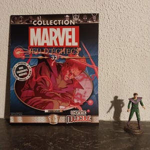 May include: A Marvel comic book titled "Collection Marvel Jeu D'Échecs 32" featuring a red and purple illustration of a character. A small figurine of a character in green and purple stands nearby.