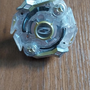 May include: A clear and gold-colored toy with a circular design. The toy has a transparent outer layer with gold accents and a central circular emblem. It is designed for play and competition.