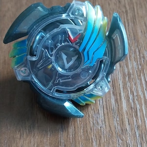 May include: A dark teal and clear plastic spinning top toy with blue and yellow accents. The toy has a V-shaped design in the center and is on a wooden surface.