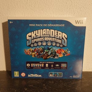 Peut inclure: Une boîte bleue pour le jeu Wii Skylanders Spyro's Adventure. La boîte présente le logo et les personnages du jeu, avec le texte "Mini Pack de Démarrage" et "Activision". La boîte est marquée d'une classification d'âge "6+" et d'une classification "7".