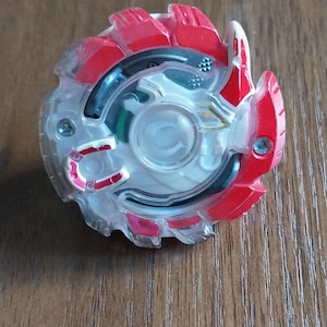 May include: A clear and red plastic toy with a circular design. The toy has a transparent center with a white circular piece in the middle. Red and white accents are on the outer edges.