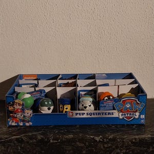Op de afbeelding: Een blauwe displaydoos met Paw Patrol Pup Squirters speelgoed. De doos heeft afbeeldingen van de personages. Elk compartiment bevat een ander personage, zoals Chase, Marshall en Skye. De doos staat op een donkere ondergrond.