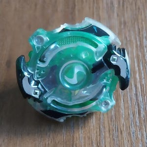 May include: A green and black plastic toy with a circular design. The toy has a central green core with a stylized 'S' shape and is surrounded by a transparent green outer ring. The edges feature black accents.