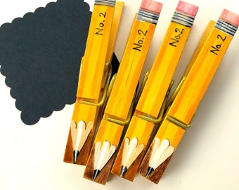 May include: Four yellow wooden clothespins painted to look like pencils with a pink eraser on top. Each clothespin has the text "No. 2" written on it.