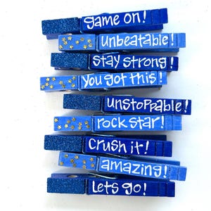 May include: A collection of blue wooden clothespins with glittered ends and white lettering. Each pin has a different motivational phrase, including "Game On!", "Unbeatable!", and "Stay Strong!" Some pins feature gold star accents.