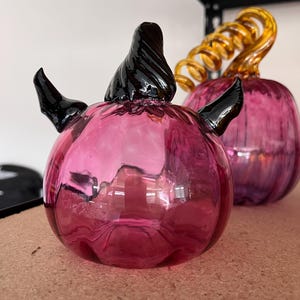 May include: Two decorative glass pumpkins. The front pumpkin is a vibrant pink with black accents on the stem and sides. The second pumpkin is a similar pink with a swirled, golden-yellow stem. Both are sitting on a light brown surface.
