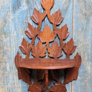 May include: A wooden wall shelf with a leaf design. The shelf is made of dark brown wood and features carved leaves around the edges and a small, round shelf in the center. The shelf is mounted on a blue and white wooden background.