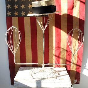 May include: A vintage hat stand with three wire hat forms. The stand is made of metal and has a white painted base. A straw hat with a black band is sitting on the top form. The stand is in front of an American flag.