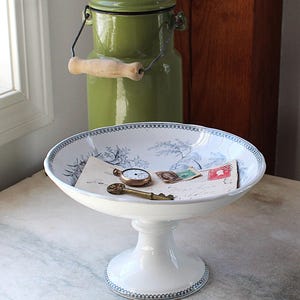 May include: A white pedestal bowl with a blue floral design and a decorative rim holds a vintage pocket watch, key, and stamps. A green metal milk can with a wooden handle sits in the background.