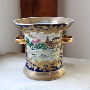 May include: A decorative ceramic vase with a flared top and two gold handles. The vase features a detailed landscape scene with Dalmatian dogs, buildings, and trees, framed by gold and navy blue accents. The base and rim are also trimmed in gold.