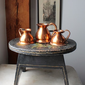 May include: Three polished copper pitchers with rounded bodies, flared rims, and curved handles. The set includes one larger central pitcher flanked by two smaller, identical pitchers, all featuring a smooth, reflective metallic finish.