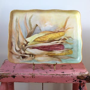 May include: Rectangular serving tray with a hand-painted corn on the cob design. The tray features a gold trim and a slightly scalloped edge. The artwork depicts yellow and red corn with green and tan husks, set against a soft, pastel background.