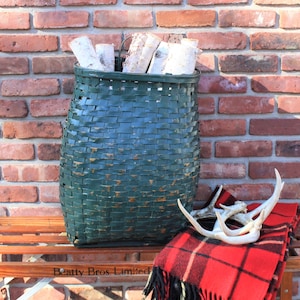 May include: A green woven basket filled with birch logs sits on a wooden bench with a red and black plaid blanket and white antlers. The basket has a metal handle and is labeled "Beatty Bros Limited".
