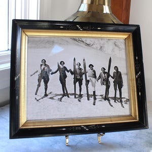 May include: Black framed vintage photograph of a group of people holding skis, standing in the snow. The frame has a gold inner border and decorative accents. The photograph is displayed on a small easel.