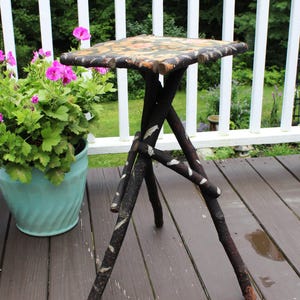 May include: A small side table featuring a square top with a floral pattern and a base constructed from three dark brown, natural tree branches. The branches have patches of white bark and are joined together to form a tripod-style support structure.