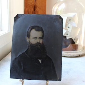 May include: A rectangular tintype photograph featuring a portrait of a person with a full dark beard and mustache, wearing a dark jacket with a visible white collar. The image has a weathered, dark gray background and is displayed on a small metal stand.
