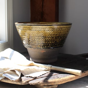 May include: A large, handcrafted ceramic bowl with a textured, chevron-like pattern in shades of brown and gold. The bowl sits on a pedestal base and is displayed on a rustic wooden surface. A linen cloth and a wooden stick are in the foreground.