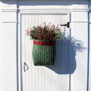 May include: A green wicker basket with a red and black plaid ribbon is hanging on a white door. The basket is filled with green branches and red berries.