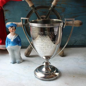 May include: A silver-toned metal trophy cup with two handles, engraved with "INVITATION REGATTA PATCHOGUE NY AUG 5TH 1913 CLASS ONE DESIGN SECOND PRIZE PRESENTED BY S.B.Y.C WON BY LADY KILLER," positioned next to a small ceramic sailor figurine.