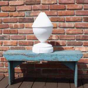 May include: A white, ceramic finial with a rounded top and a base sits on a blue, wooden bench. The bench is weathered and has a distressed finish. The finial is a decorative architectural element.
