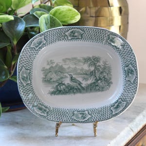 May include: Oval serving platter with a green and white landscape design. The platter features a detailed bird and mountain scene in the center, surrounded by a checkered border and decorative accents. The platter is displayed on a gold-colored stand.