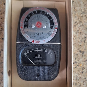 May include: A vintage General Electric exposure meter in a cardboard box. The device features a black textured casing, a circular silver dial with f-stop and light settings, and a lower rectangular window displaying light levels in footcandles.