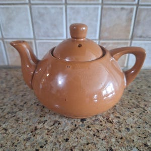 May include: A glossy, light brown ceramic teapot with a rounded body, a curved spout, a sturdy handle, and a matching lid featuring a small knob handle and a steam vent hole. The teapot has a smooth, reflective finish and a simple, functional design.