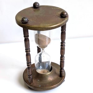 May include: An hourglass featuring a clear glass bulb filled with fine tan sand, held within a dark metallic frame. The frame consists of a circular top and base connected by four turned pillars, with small spherical feet supporting the structure.