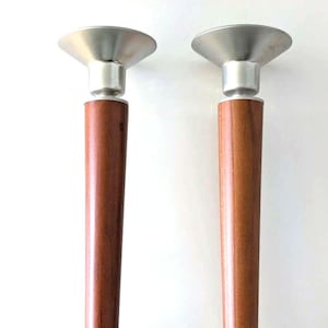 May include: A pair of candle holders featuring cylindrical brown wooden stems, silver-toned metal bobeches at the top, and circular clear glass bases. The design is vertical and symmetrical, showcasing a combination of wood, metal, and glass.