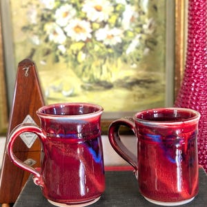 May include: Two handmade ceramic mugs with a deep red glaze and blue accents. The mugs have a rounded shape with a handle and a white rim. They are sitting on a dark surface, with a painting and decorative items in the background.