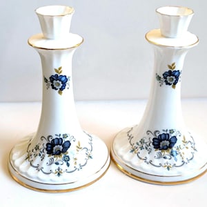 May include: A pair of white porcelain candle holders featuring a flared base and a fluted top. The surface is decorated with blue floral patterns, green leaves, and delicate grey scrollwork, finished with a thin gold trim along the edges.