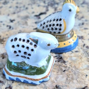 May include: Two ceramic animal figurines with perforated backs. One is a white rabbit on a green and blue base, and the other is a white bird with yellow and blue accents on a circular base. Both figures feature hand-painted details and holes.