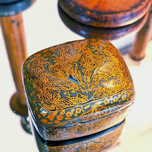 May include: A square, decorative box with a detailed, ornate design in gold, orange, and blue. The box sits on a reflective surface, with other wooden objects in the background.