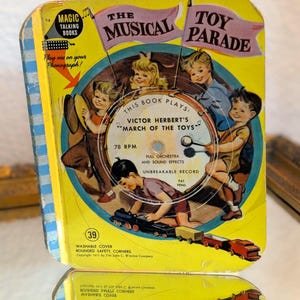 May include: Vintage children's book titled "The Musical Toy Parade." The cover features illustrations of children, musical instruments, and toy trains. The book plays "March of the Toys" by Victor Herbert. The cover is yellow with blue accents and text.