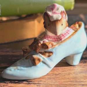 May include: A vintage ceramic figurine featuring a mouse wearing a chef's hat and scarf, seated in a light blue high-heeled shoe. The shoe has brown floral accents. The figurine is set on a wooden surface.