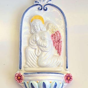 May include: A ceramic wall-mounted holy water font featuring a white kneeling angel with a yellow halo and pink wings. The arched frame is decorated with blue trim and a brown leather hanging cord. The basin is adorned with two small pink flowers.